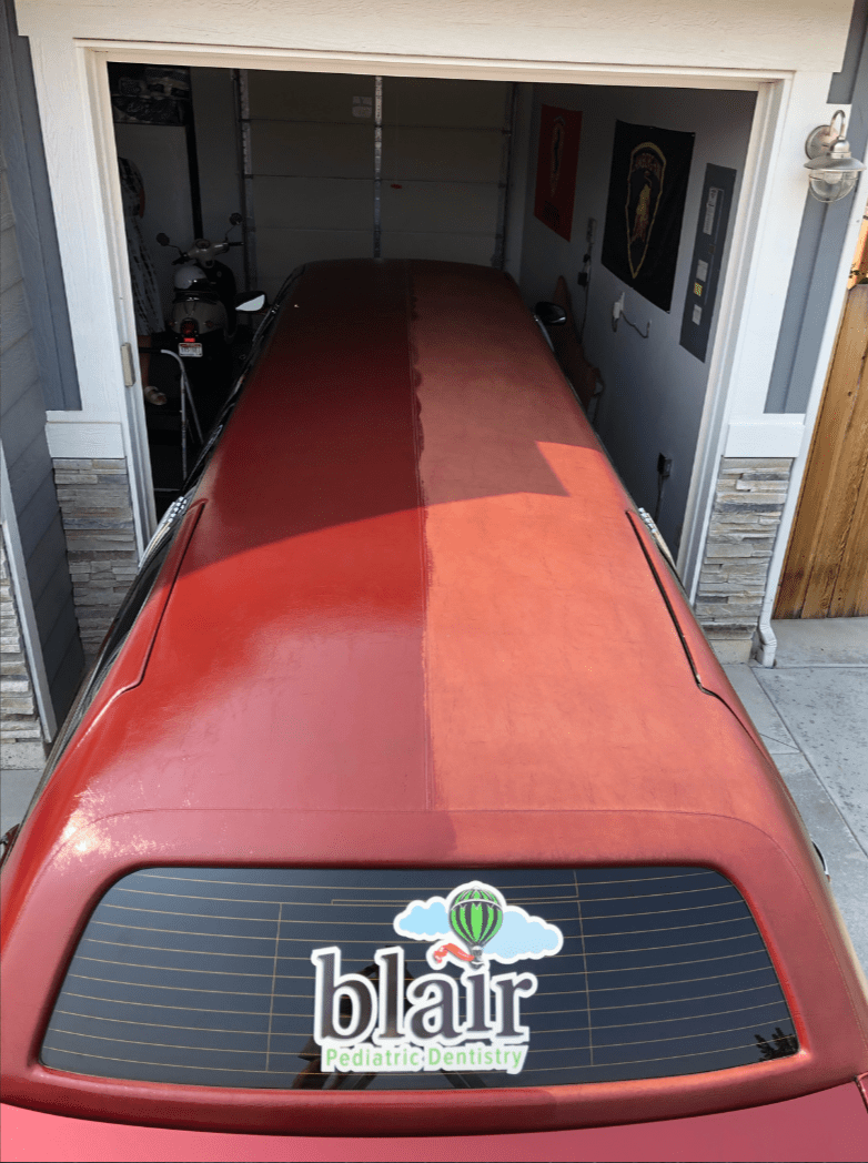 Top view of a red stretch limousine parked in a residential garage, showing a clear contrast between a freshly cleaned or restored left side and a dull, oxidized right side.