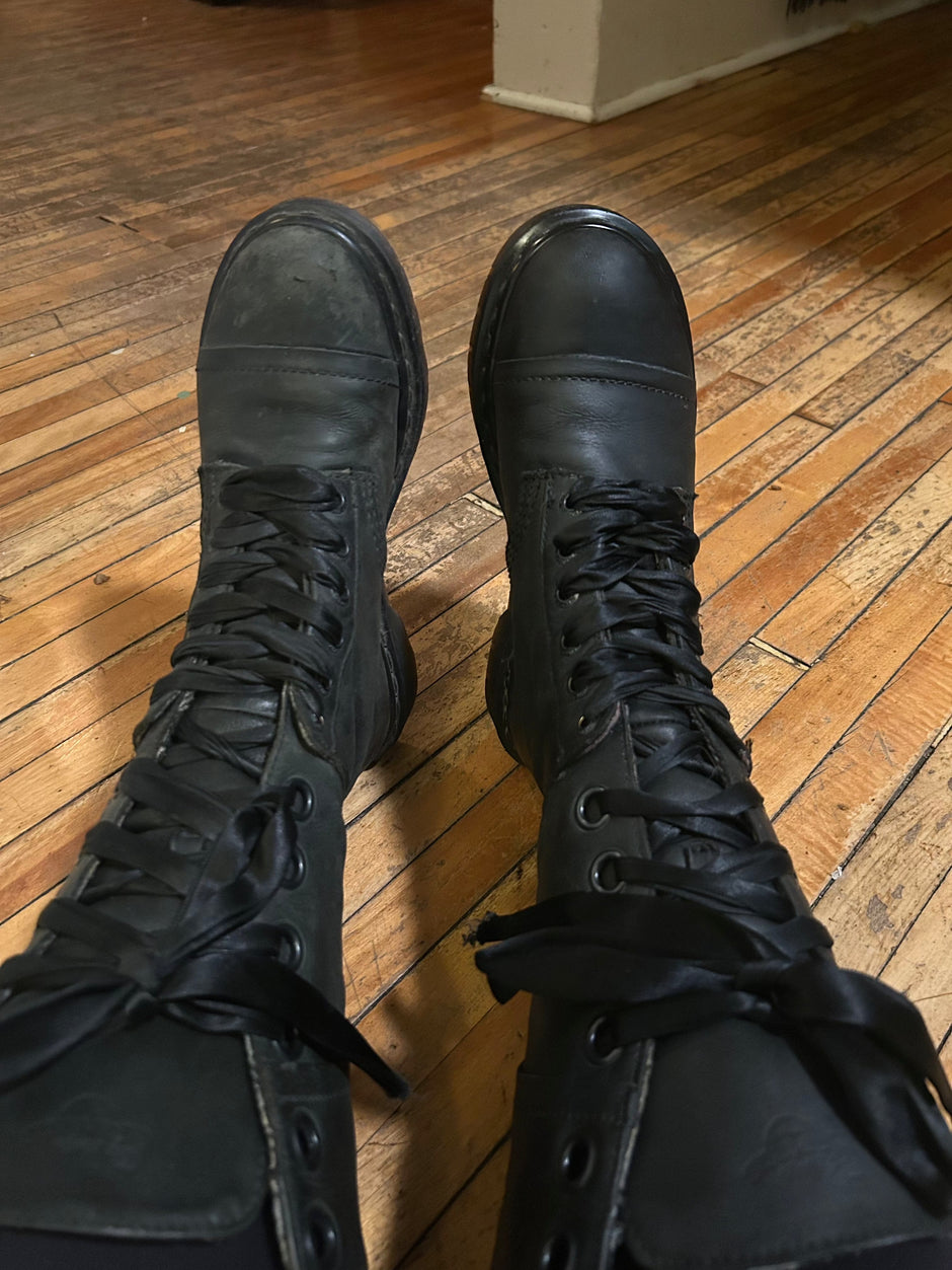 Overhead view of a person wearing black leather lace-up Dr. Martens boots on a hardwood floor. The left boot shows signs of scuffing and dullness, while the right boot appears freshly restored or polished, with a smoother and more lustrous finish. The image highlights the contrast and effectiveness of Leather Better conditioner.