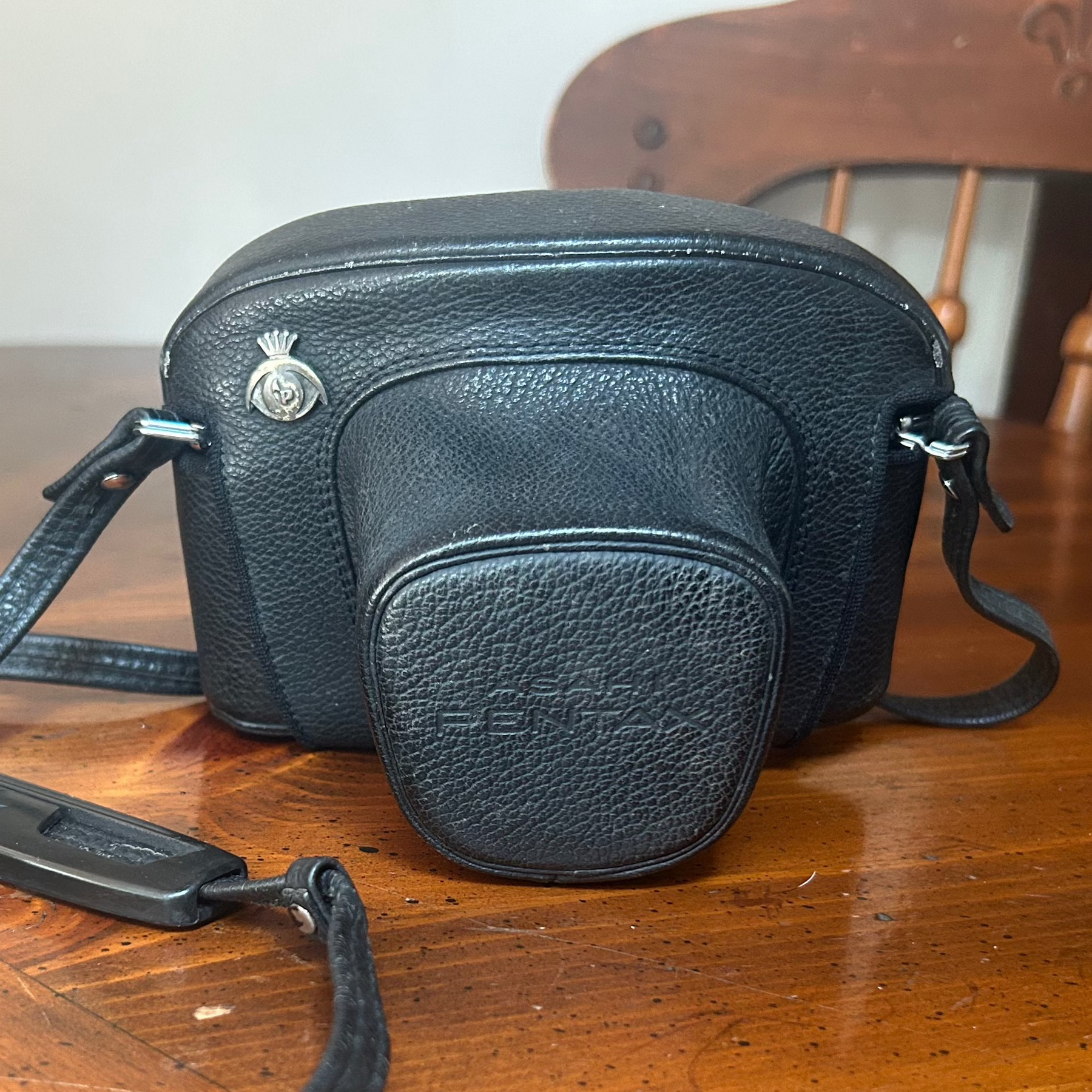 Black leather Pentax camera case shown on a wooden table, with a slightly dull and uneven surface. The leather has a matte appearance and lacks vibrancy, showing signs of age or dryness.