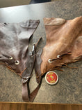 Load image into Gallery viewer, A pair of riding chaps laying on a table. The left leather side has been treated with Leather better, and the right side is hasn't been treated yet.  The difference in colour is dramatic with the treated side being a rich chocolate colour and the untreated side being closer to tan. A can of Leather better is visible beside the chaps.