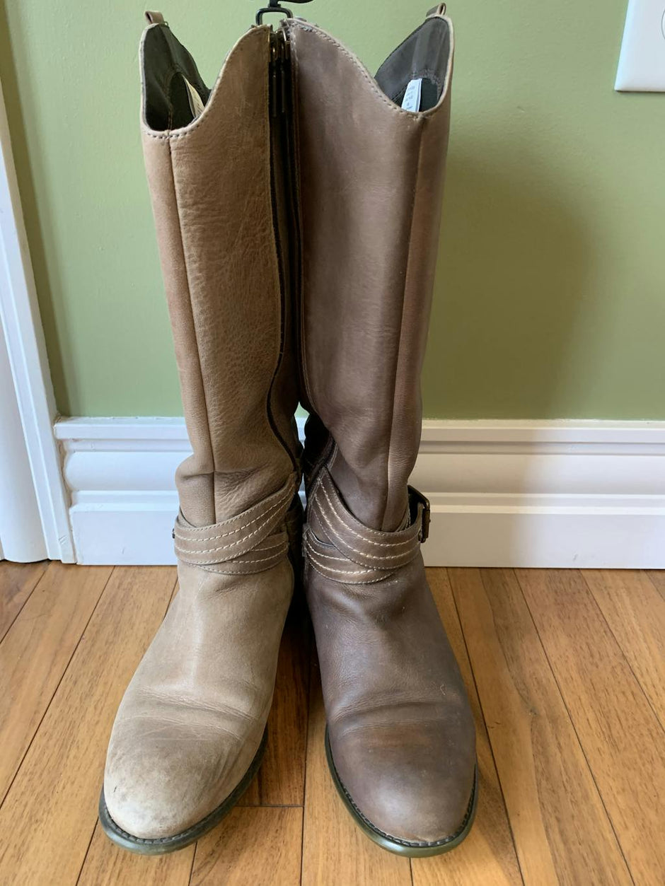 Side-by-side comparison of a pair of tall brown leather boots, showing one boot restored to a rich, even tone while the other remains faded and scuffed. The image highlights the dramatic results of leather conditioning and restoration on footwear.