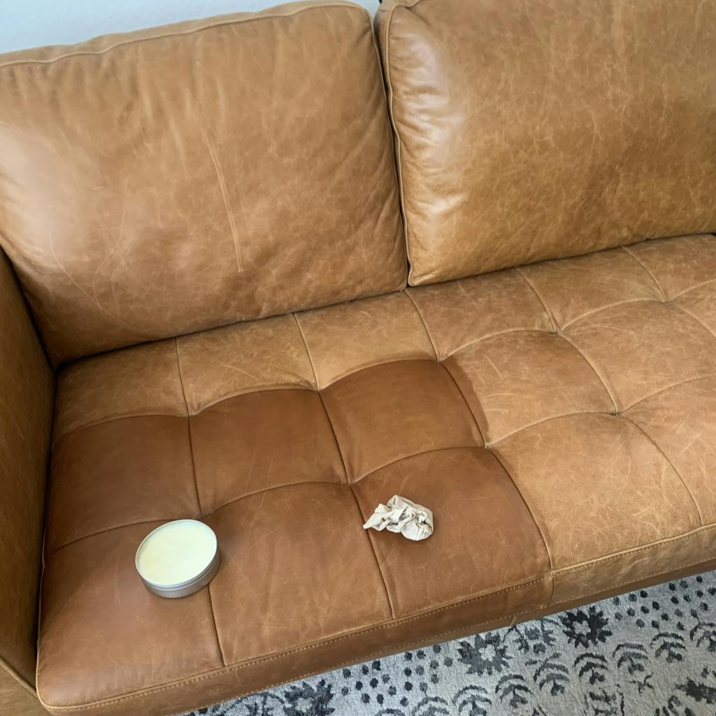 A tan faded leather couch with an open can of Leather better with a rag on top of a treated patch of the faded couch.