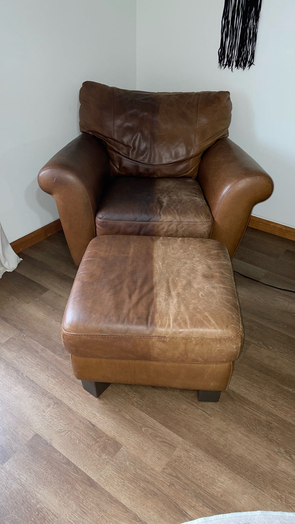 Brown leather armchair and matching ottoman in a corner of a room, with the ottoman showing a clear split between a conditioned and untreated side. The right side of the ottoman appears darker and more supple after leather restoration, while the left side remains dry and faded, demonstrating a visible before-and-after contrast.