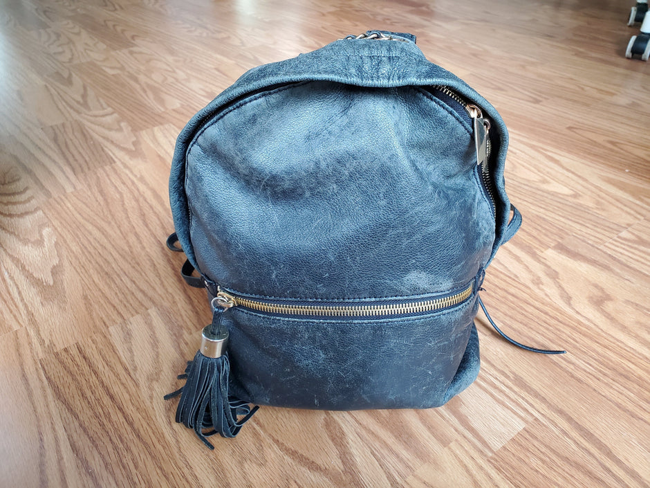 Faded blue leather backpack with visible surface wear, discolouration, and dull patches across the front and top. The leather appears dry and scuffed, especially near the zipper and seams.