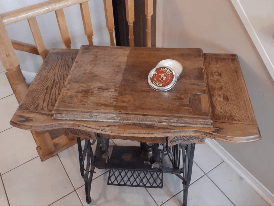 Sewing machine desk on a tile floor with iron legs. Right side of the desk has been treated with Leather better, and the left side untreated to showcase the results. A can of Leather better sits on open on the desk.