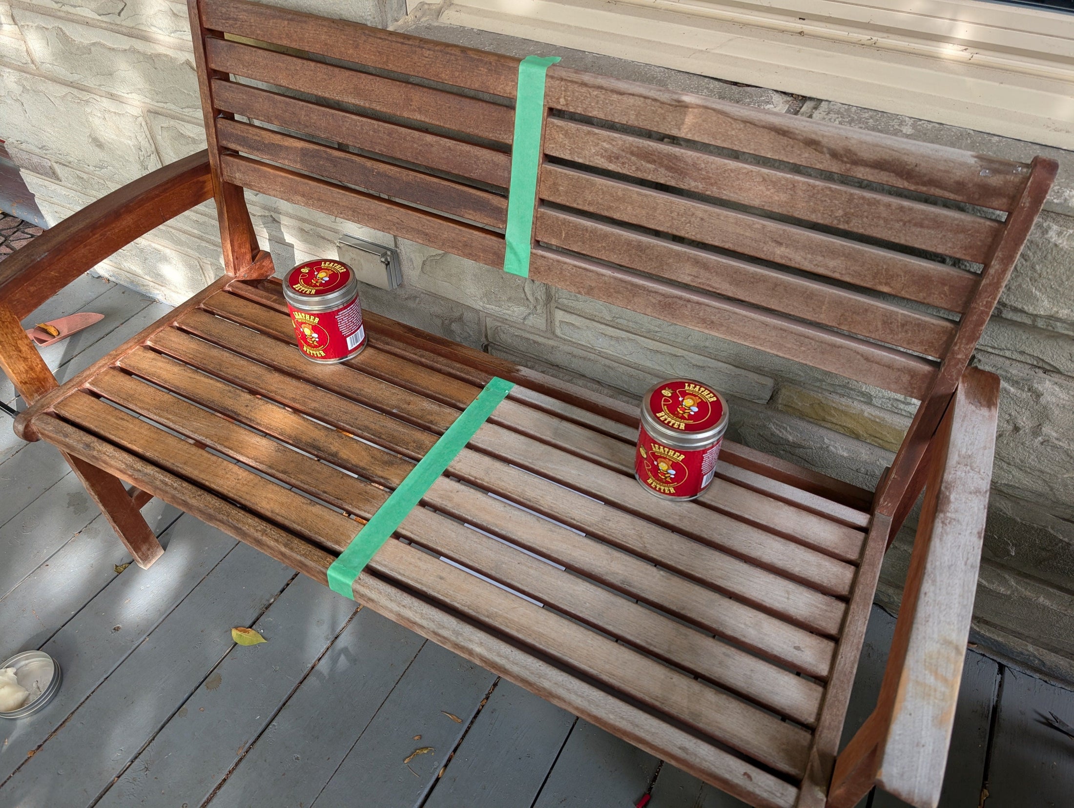 A wooden porch bench is getting a makeover with Leather Better. The bench is taped down the middle. The left side is worn, faded and dull looking whereas the right side has been treated and the colour and texture has been restored. 