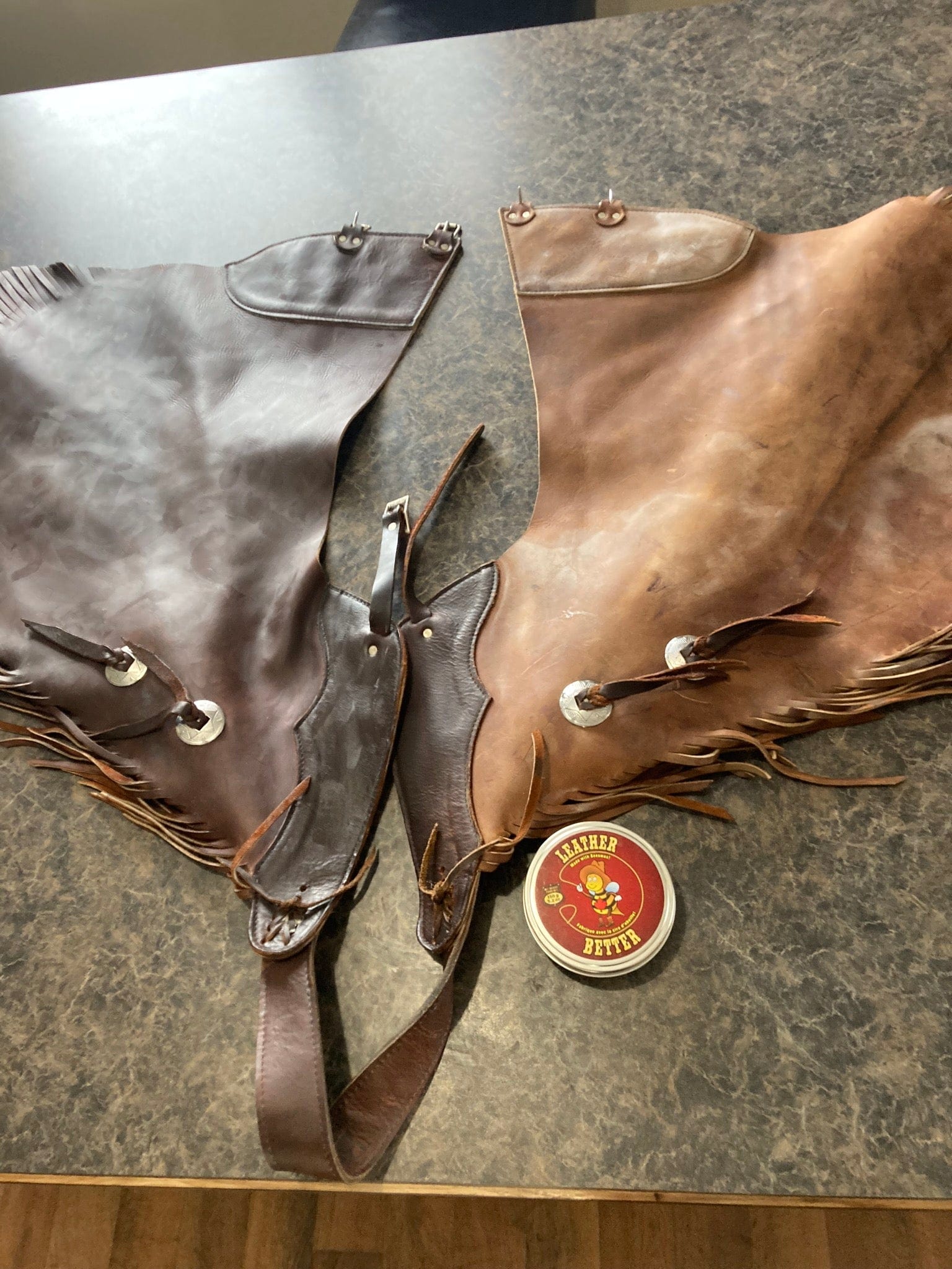A pair of riding chaps laying on a table. The left leather side has been treated with Leather better, and the right side is hasn't been treated yet.  The difference in colour is dramatic with the treated side being a rich chocolate colour and the untreated side being closer to tan. A can of Leather better is visible beside the chaps.